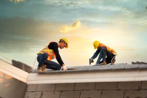 Roofing workers inspect exposed roof decking beside stacked shingles on a residential home.