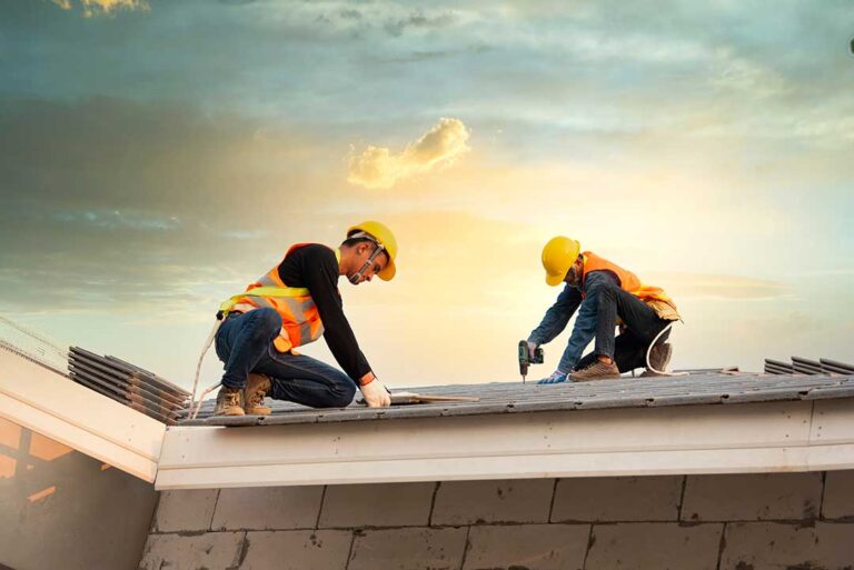 Roofing workers inspect exposed roof decking beside stacked shingles on a residential home.
