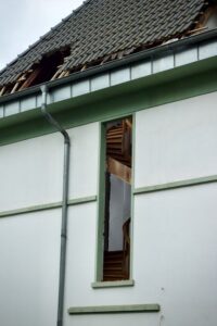 A homeowner examines a ceiling water stain beneath a roof leak while damp insulation is visible in the attic.