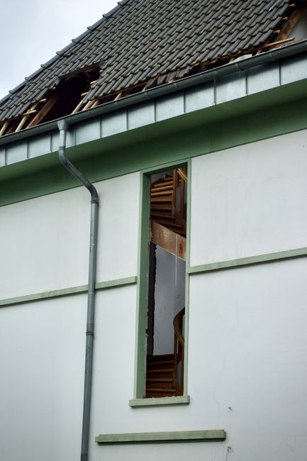 A homeowner examines a ceiling water stain beneath a roof leak while damp insulation is visible in the attic.